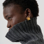A close-up side profile of a person with short dark hair wearing a dark gray ribbed turtleneck sweater and a small gold square stud earring with a tiny white monogram.