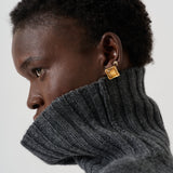 A close-up side profile of a person with short dark hair wearing a dark gray ribbed turtleneck sweater and a small gold square stud earring with a tiny white monogram.