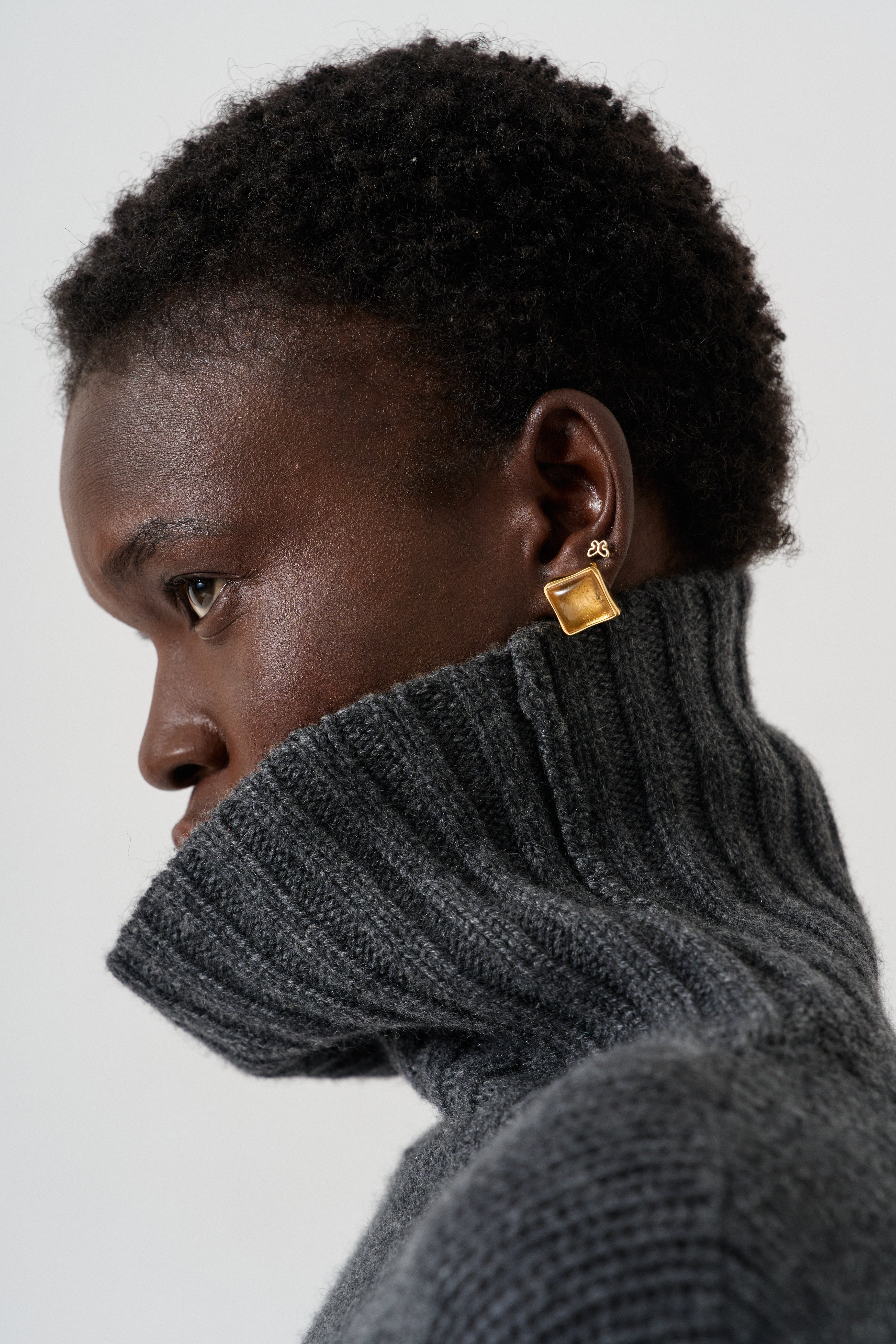 A close-up side profile of a person with short dark hair wearing a dark gray ribbed turtleneck sweater and a small gold square stud earring with a tiny white monogram.
