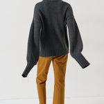 A model shown from the back wearing a dark gray oversized turtleneck sweater, mustard yellow trousers, and brown slippers on a white studio background.