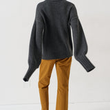 A model shown from the back wearing a dark gray oversized turtleneck sweater, mustard yellow trousers, and brown slippers on a white studio background.