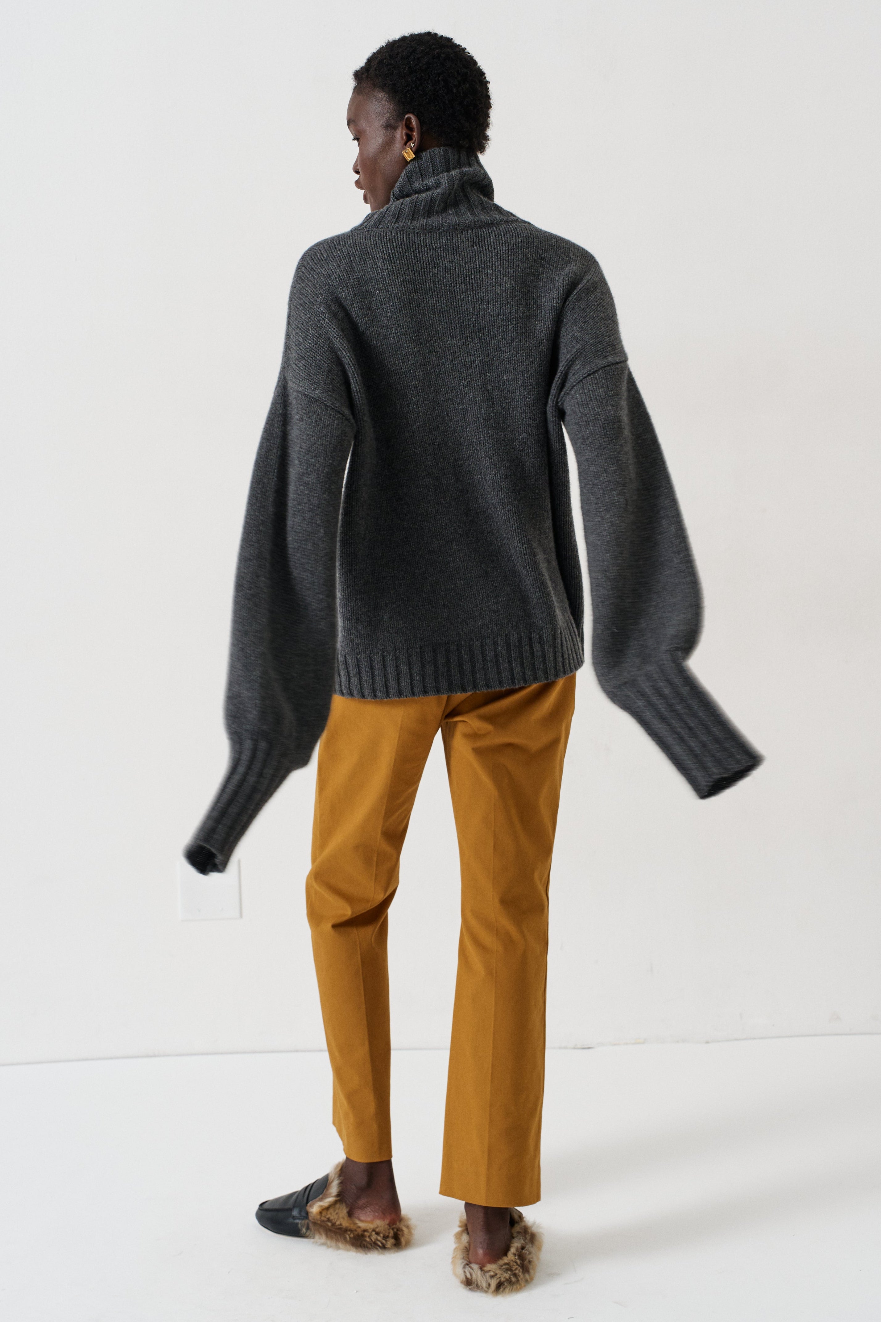 A model shown from the back wearing a dark gray oversized turtleneck sweater, mustard yellow trousers, and brown slippers on a white studio background.