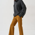 A model wearing a dark gray turtleneck sweater, mustard yellow wide-leg trousers, and black loafers with fur trim, standing against a plain white studio background.