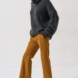 A model wearing a dark gray turtleneck sweater, mustard yellow wide-leg trousers, and black loafers with fur trim, standing against a plain white studio background.