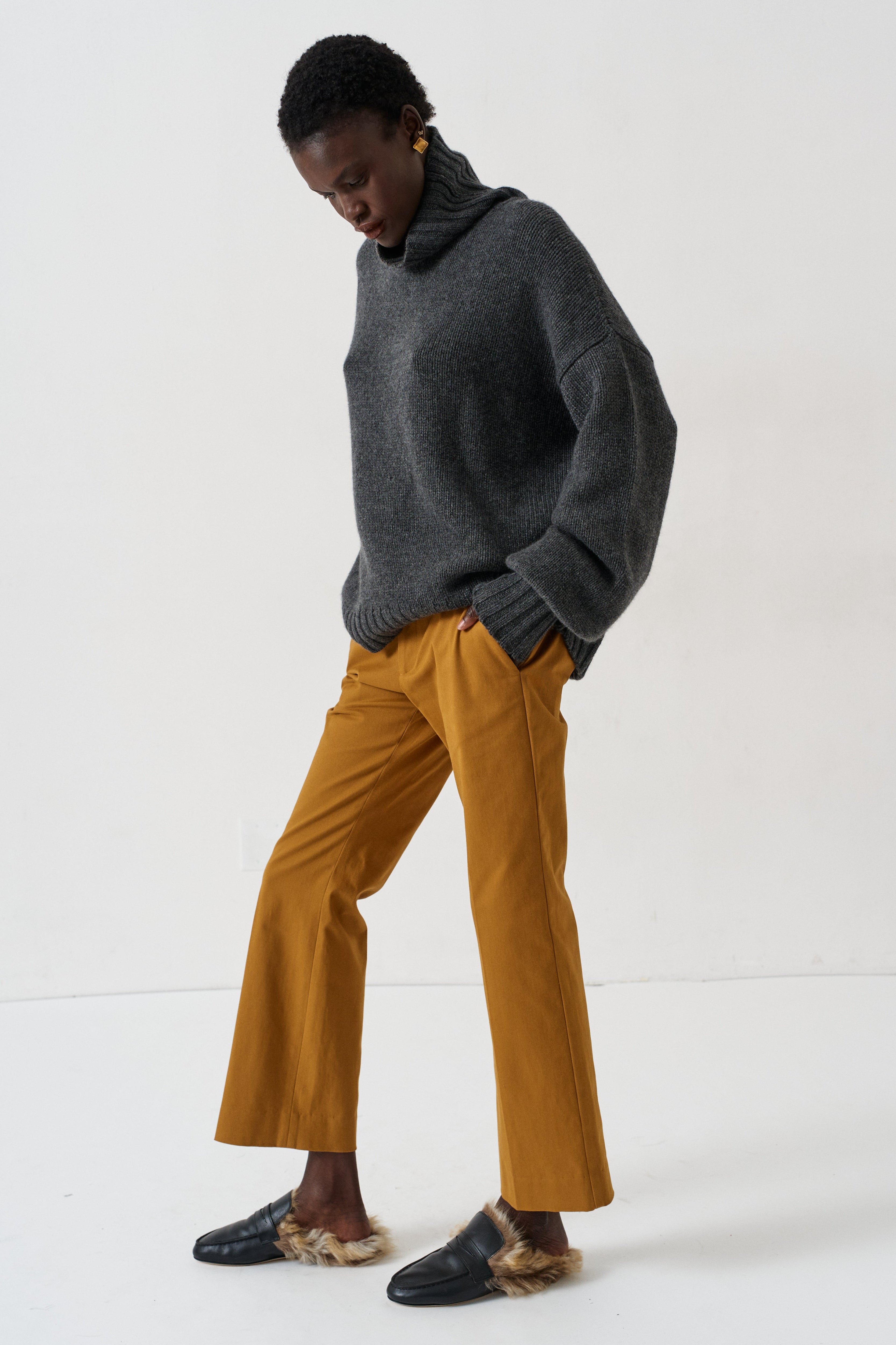 A model wearing a dark gray turtleneck sweater, mustard yellow wide-leg trousers, and black loafers with fur trim, standing against a plain white studio background.