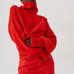 A model wearing a vibrant red oversized turtleneck sweater and matching red trousers, posed against a clean white studio background.