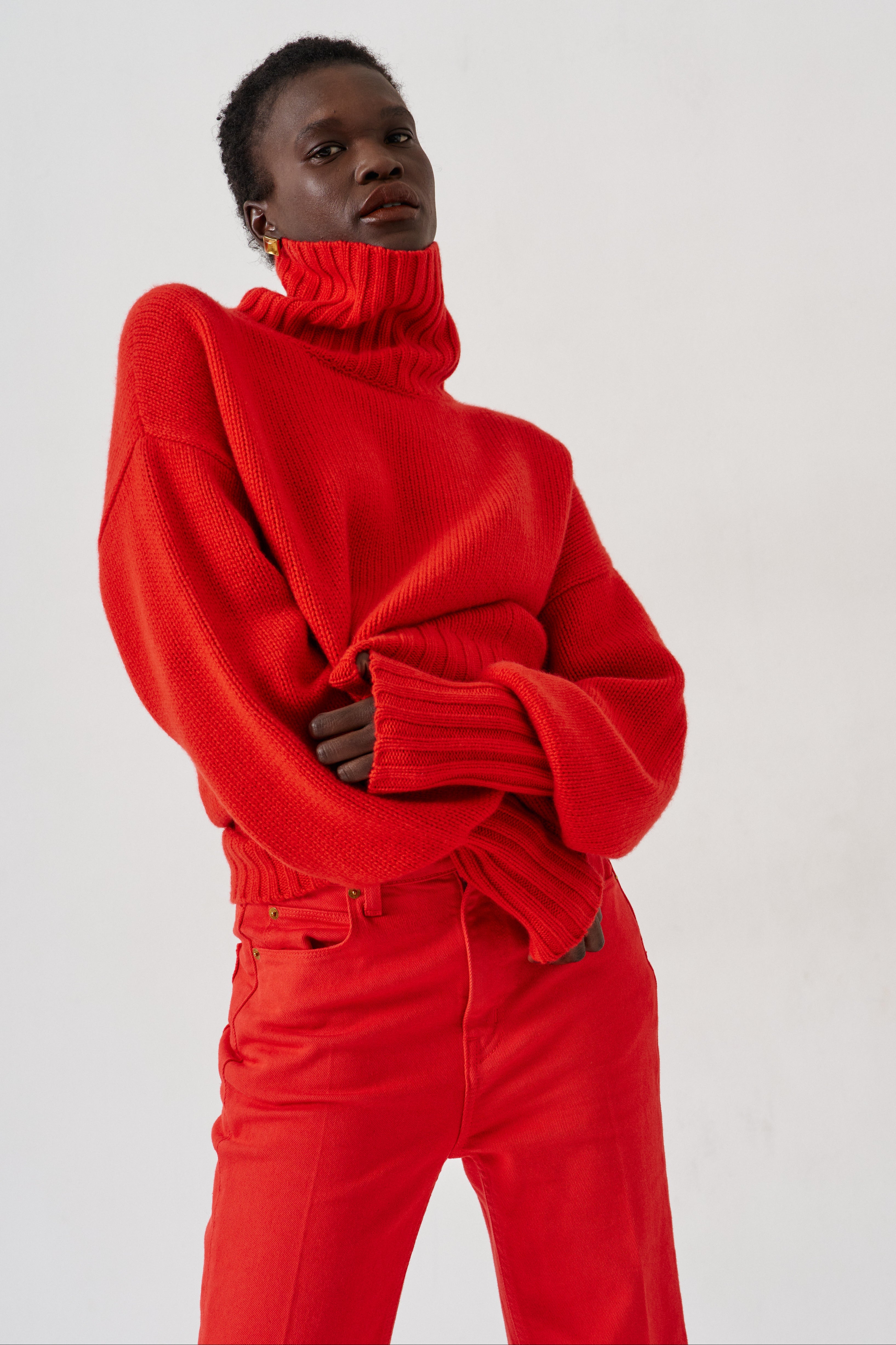 A model wearing a vibrant red oversized turtleneck sweater and matching red trousers, posed against a clean white studio background.