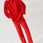 A model wearing a bright red knit turtleneck sweater and matching red trousers, paired with brown suede loafers, standing against a plain white studio background.