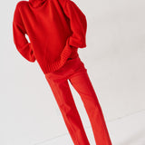 A model wearing a bright red knit turtleneck sweater and matching red trousers, paired with brown suede loafers, standing against a plain white studio background.