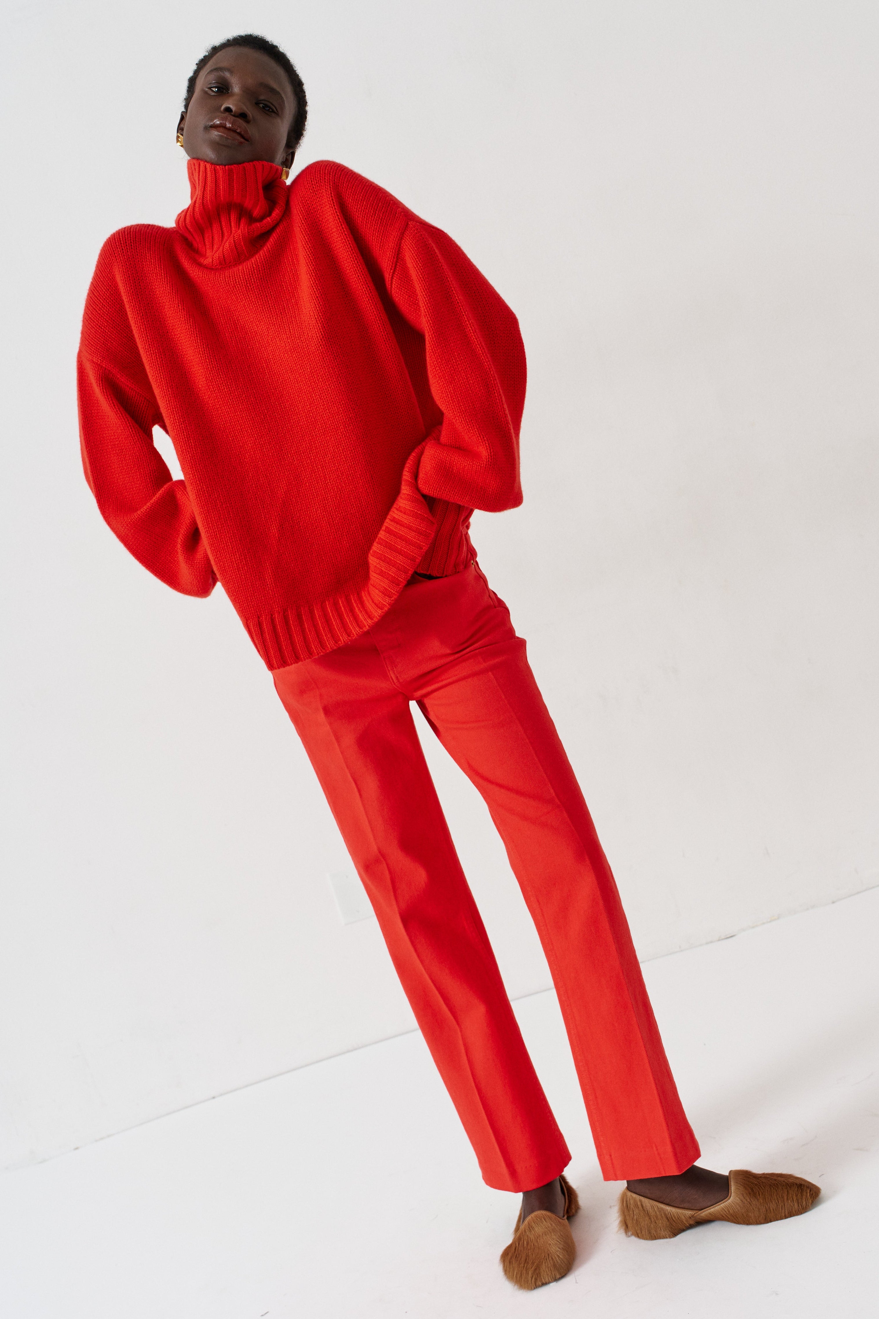 A model wearing a bright red knit turtleneck sweater and matching red trousers, paired with brown suede loafers, standing against a plain white studio background.