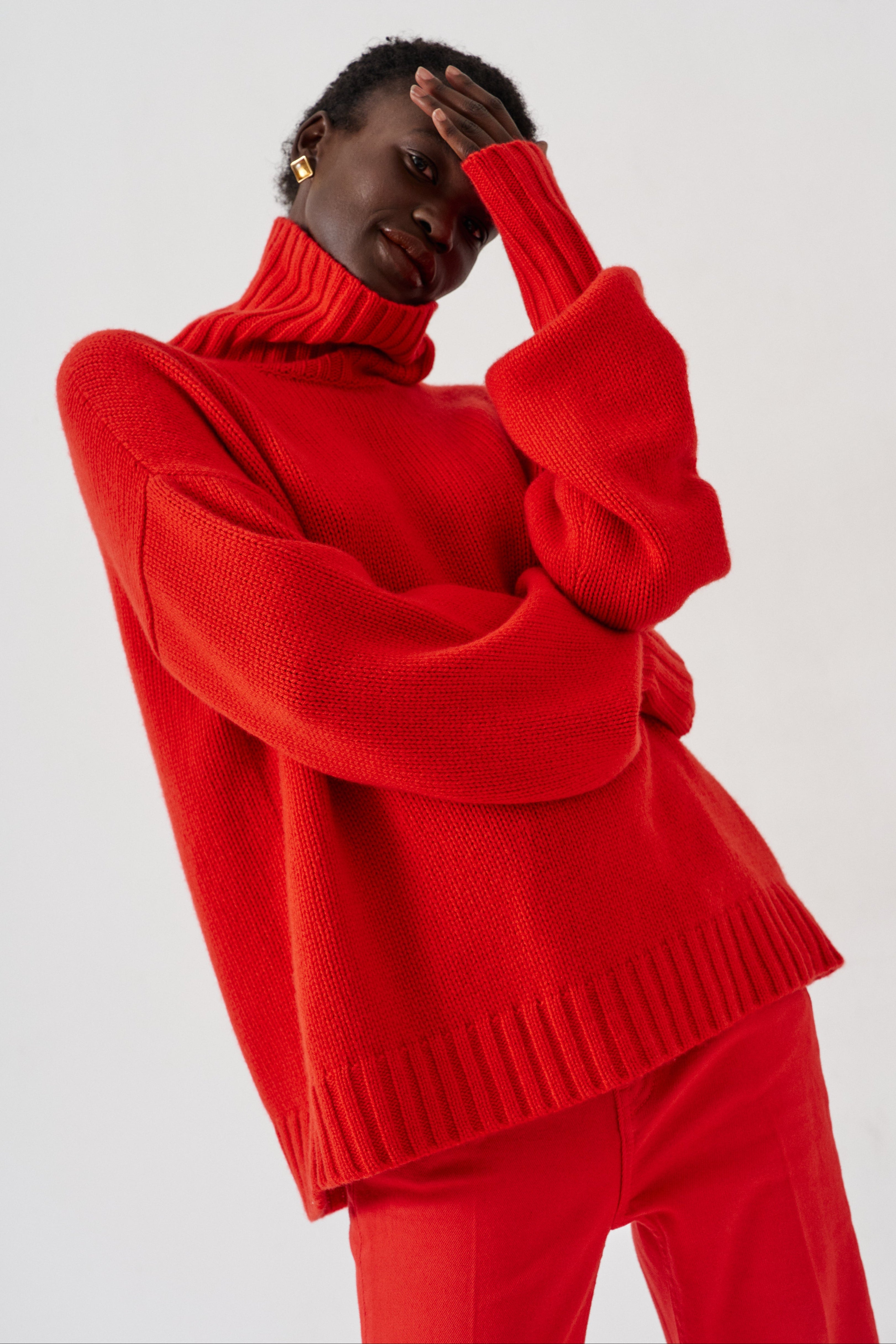 A model wearing a vibrant red knit turtleneck sweater and matching red trousers, posing against a light gray studio background.