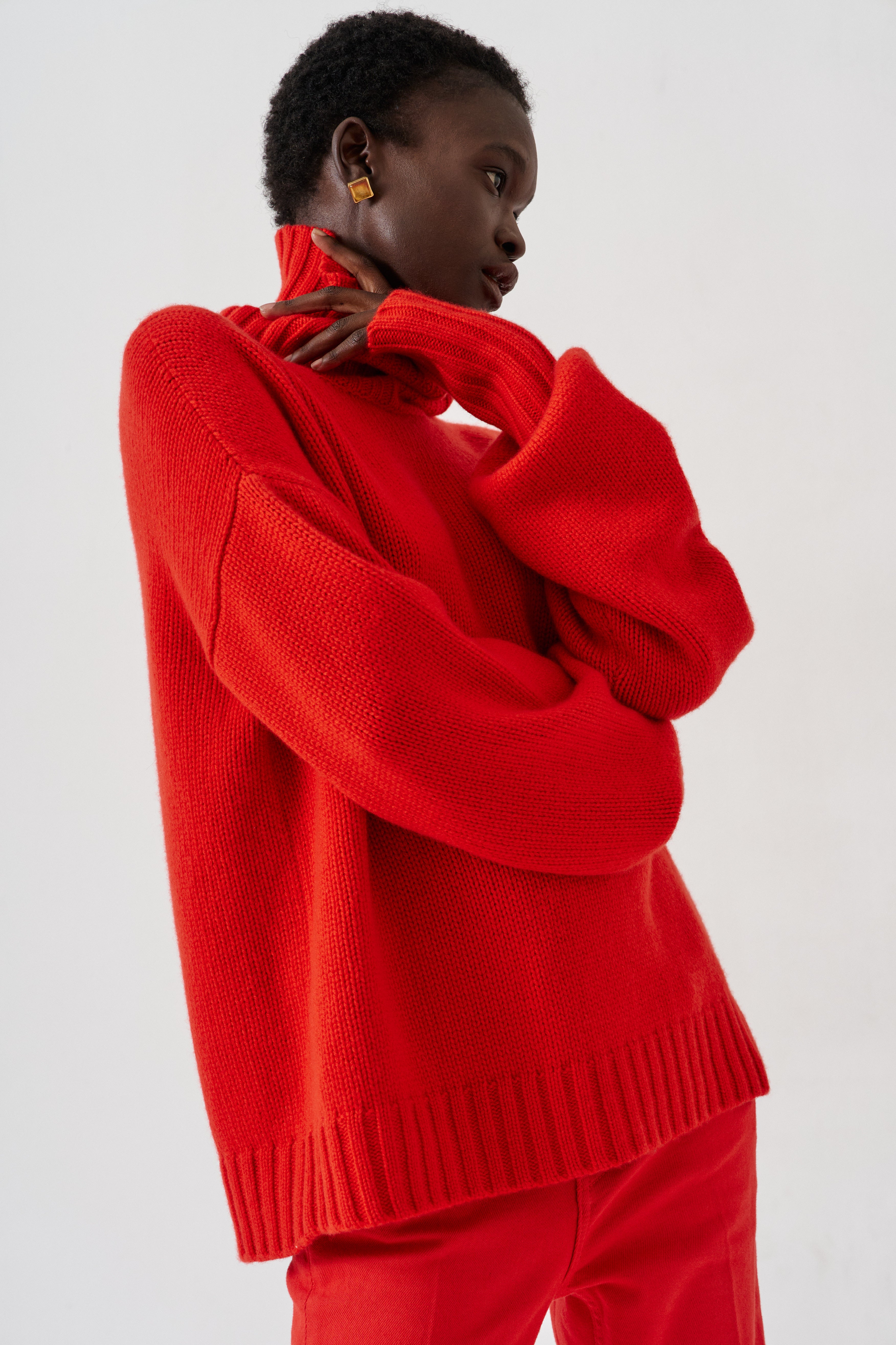 A model wearing a bright red knit sweater and matching red trousers, shown from the side against a plain light gray studio background.