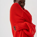 A model wearing a vibrant red chunky knit sweater, wrapped around their body, photographed against a clean white studio background.