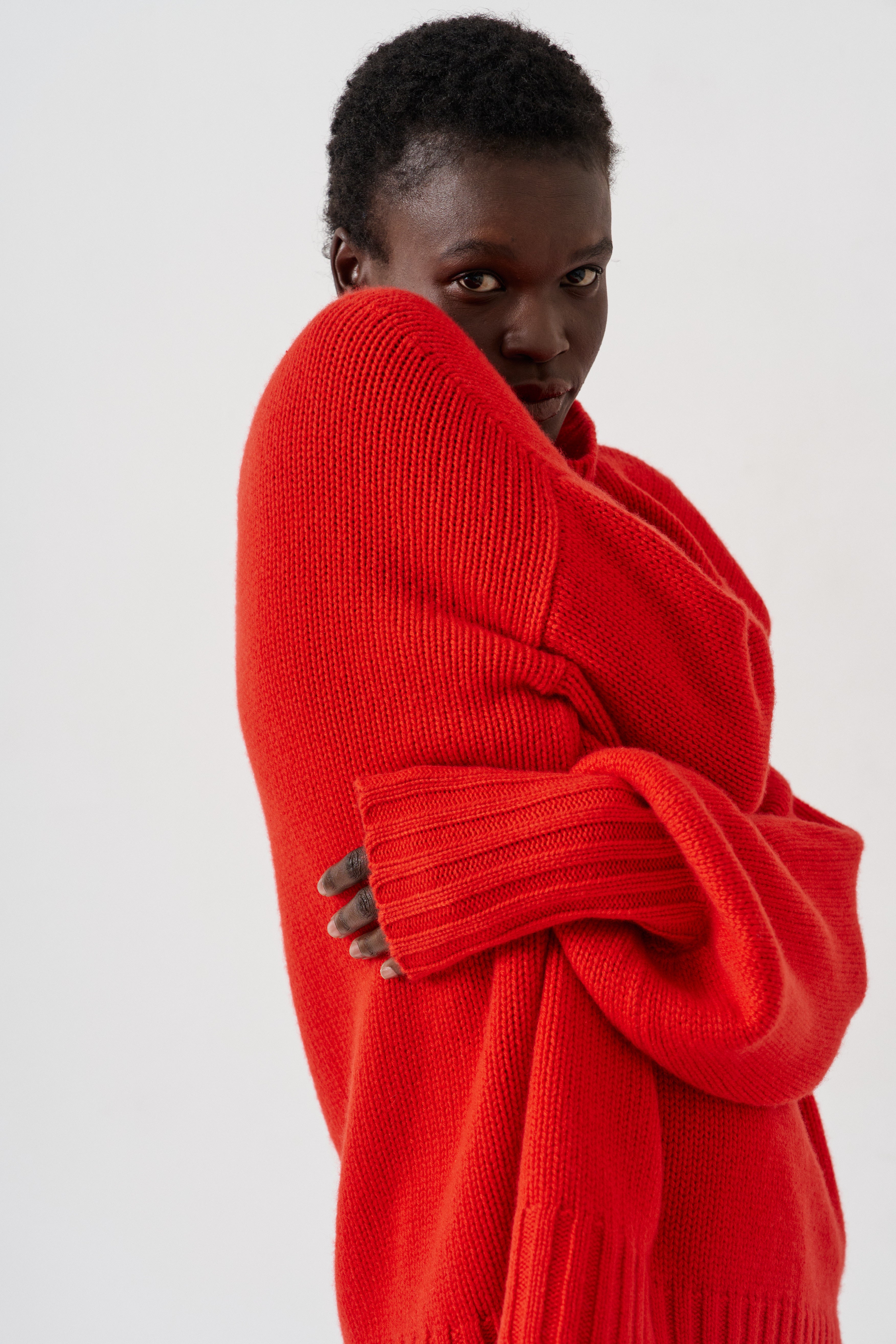 A model wearing a vibrant red chunky knit sweater, wrapped around their body, photographed against a clean white studio background.