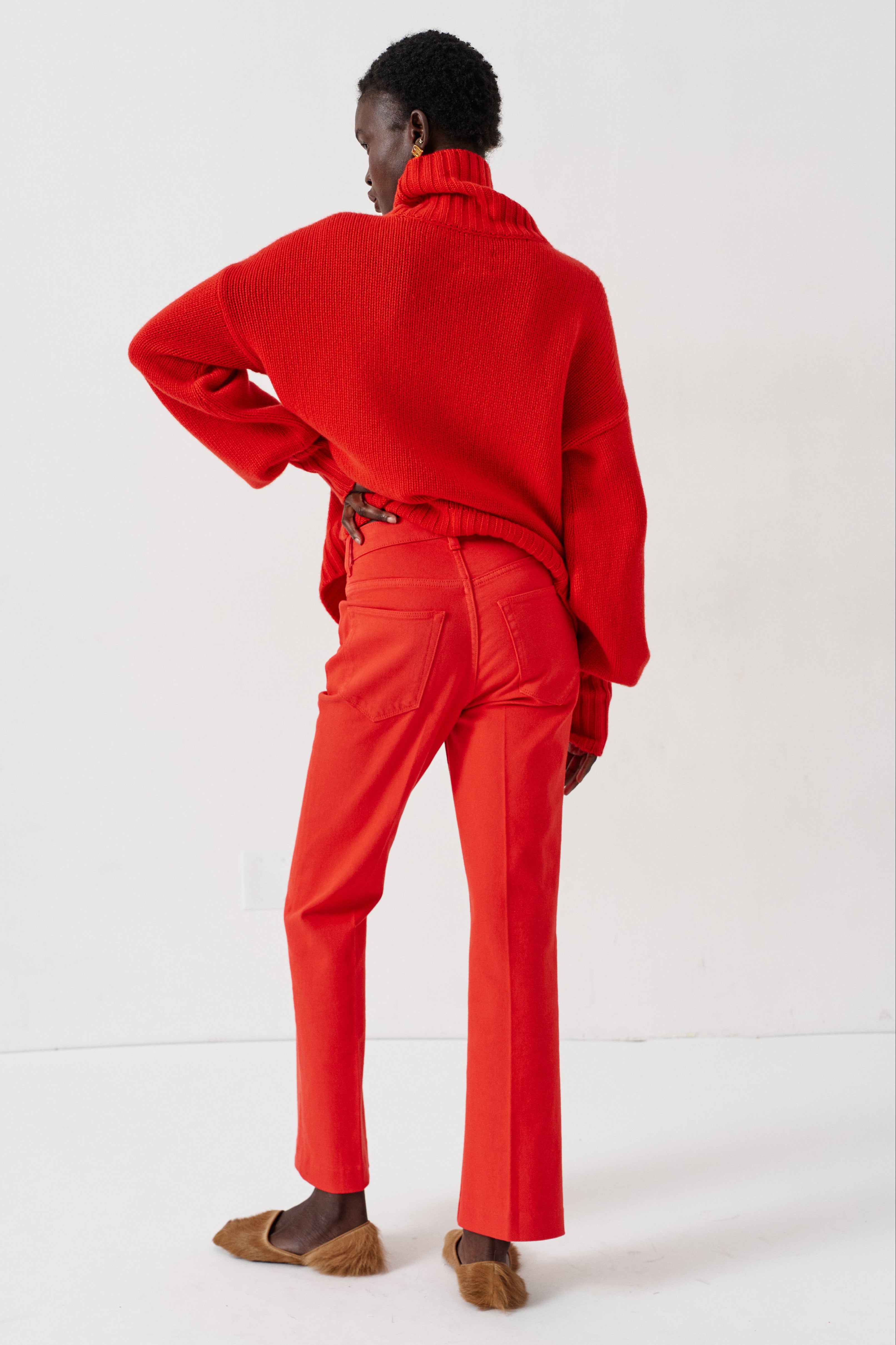 A model shown from the back wearing a bright red knit turtleneck sweater and matching red trousers, paired with brown shoes, against a clean white studio background.