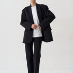 A model stands against a plain white studio background wearing a black double-breasted blazer with structured shoulders, matching black trousers, a white crewneck t-shirt, and black leather loafers with light brown fur trim.
