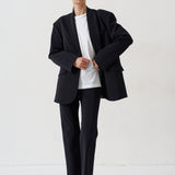 A model stands against a plain white studio background wearing a black double-breasted blazer with structured shoulders, matching black trousers, a white crewneck t-shirt, and black leather loafers with light brown fur trim.