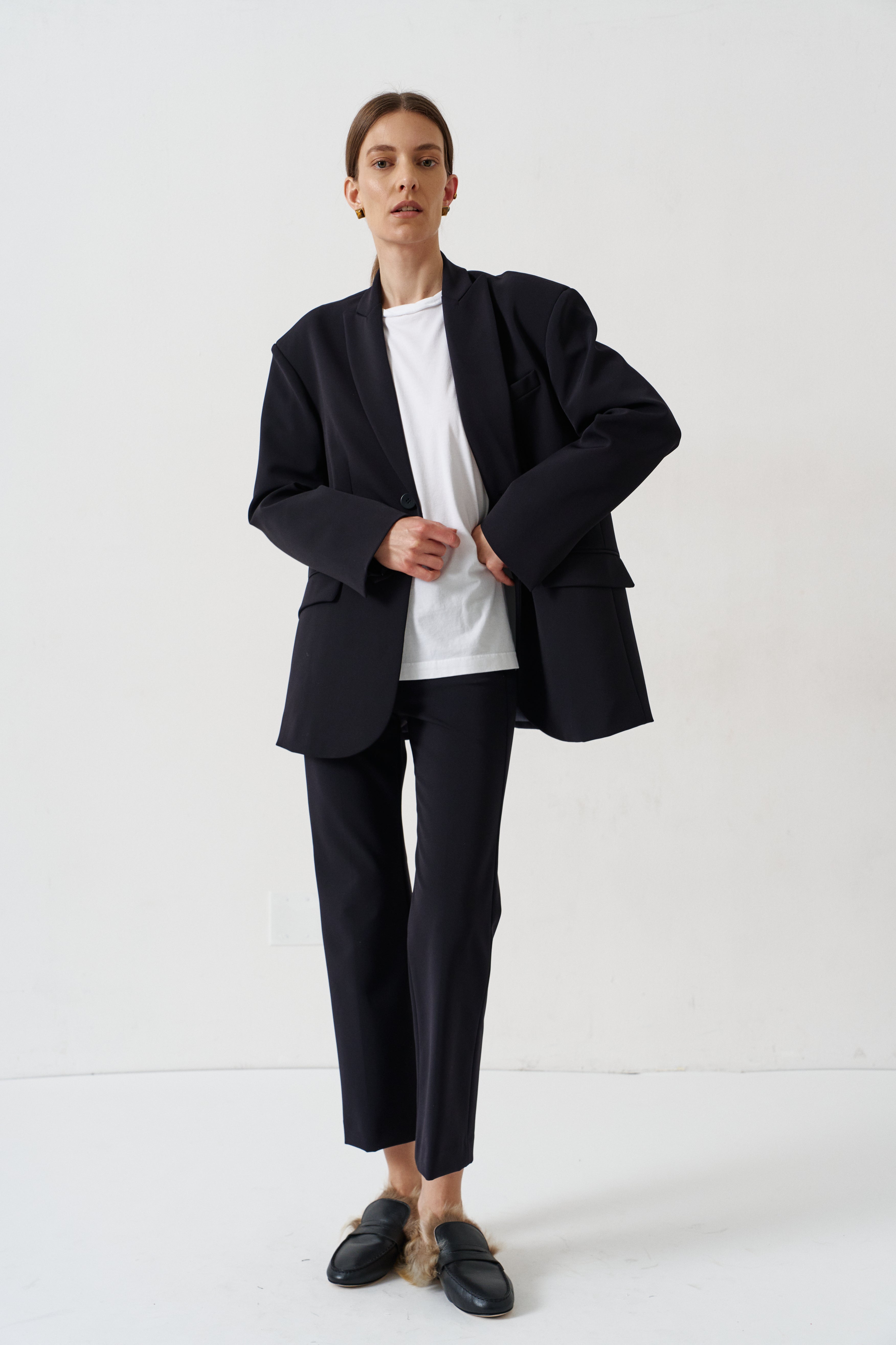 A model stands against a plain white studio background wearing a black double-breasted blazer with structured shoulders, matching black trousers, a white crewneck t-shirt, and black leather loafers with light brown fur trim.