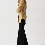 A woman stands in profile wearing a beige and black vertically striped button-up shirt, black flared trousers, and dark pointed-toe heels against a plain white studio background.