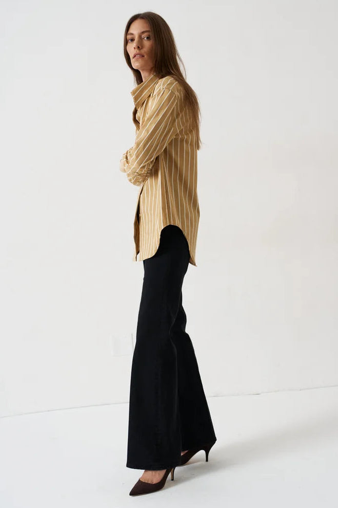 A woman stands in profile wearing a beige and black vertically striped button-up shirt, black flared trousers, and dark pointed-toe heels against a plain white studio background.