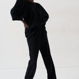 A model wearing a black two-piece lounge set consisting of a loose-fitting top with voluminous sleeves and matching wide-leg pants, styled with patterned shoes against a plain white studio background.