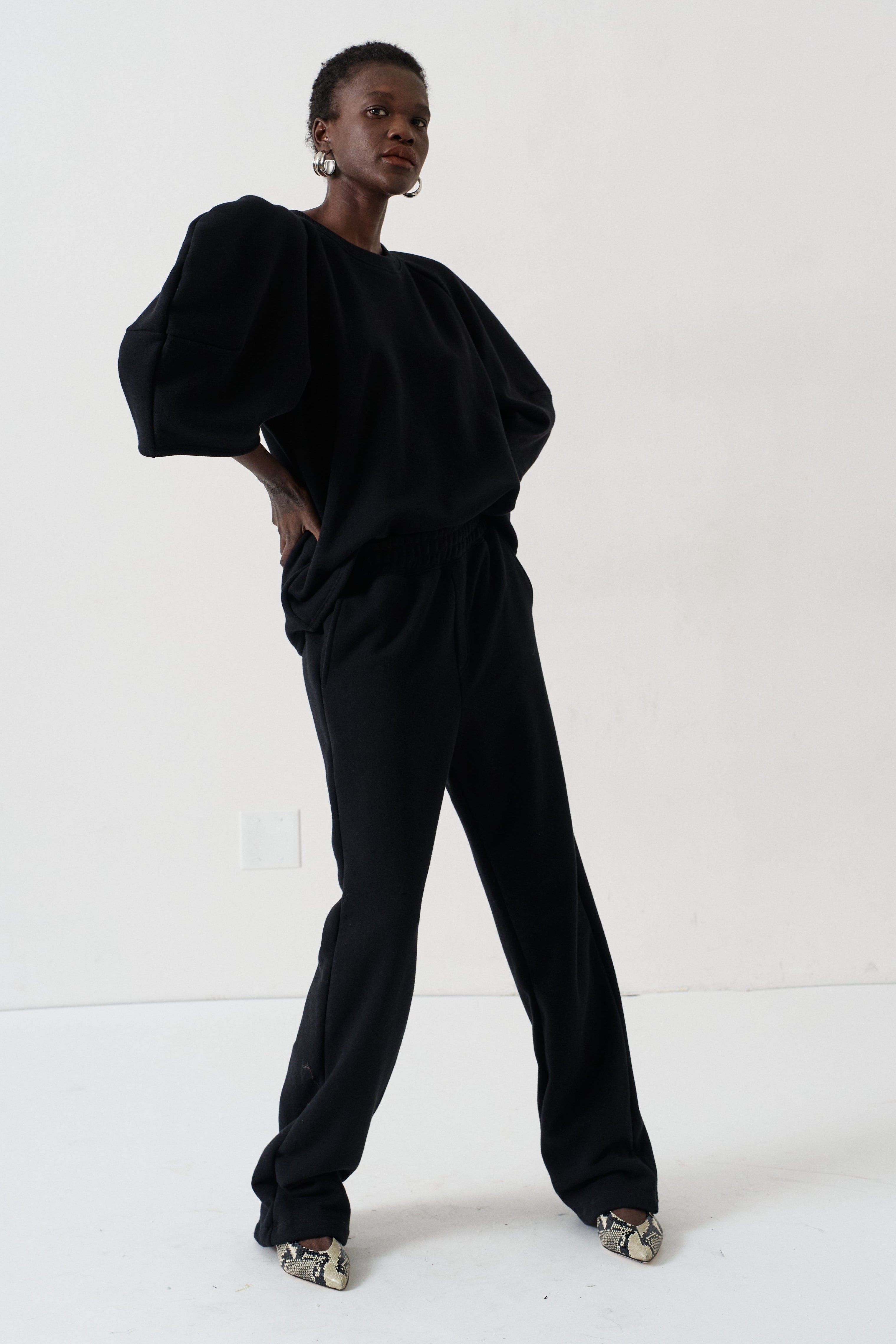 A model wearing a black two-piece lounge set consisting of a loose-fitting top with voluminous sleeves and matching wide-leg pants, styled with patterned shoes against a plain white studio background.