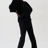 A model wearing a black two-piece lounge set consisting of a loose-fitting top with voluminous sleeves and matching wide-leg pants, styled with patterned shoes against a plain white studio background.