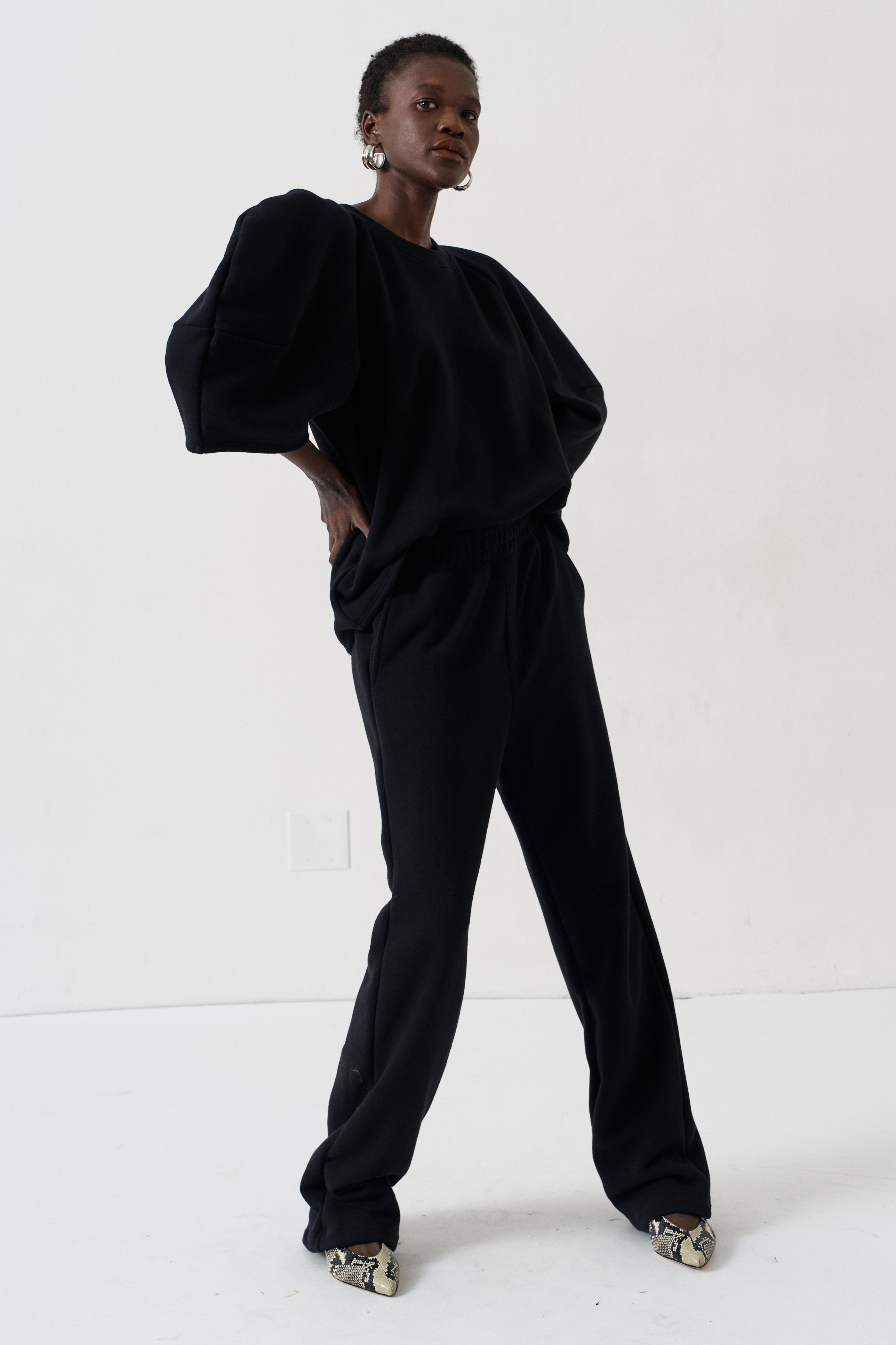 A model wearing a black two-piece lounge set consisting of a loose-fitting top with voluminous sleeves and matching wide-leg pants, styled with patterned shoes against a plain white studio background.