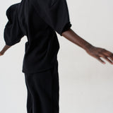 A model from behind wearing a black puff-sleeve top and matching black wide-leg trousers against a plain white studio background.