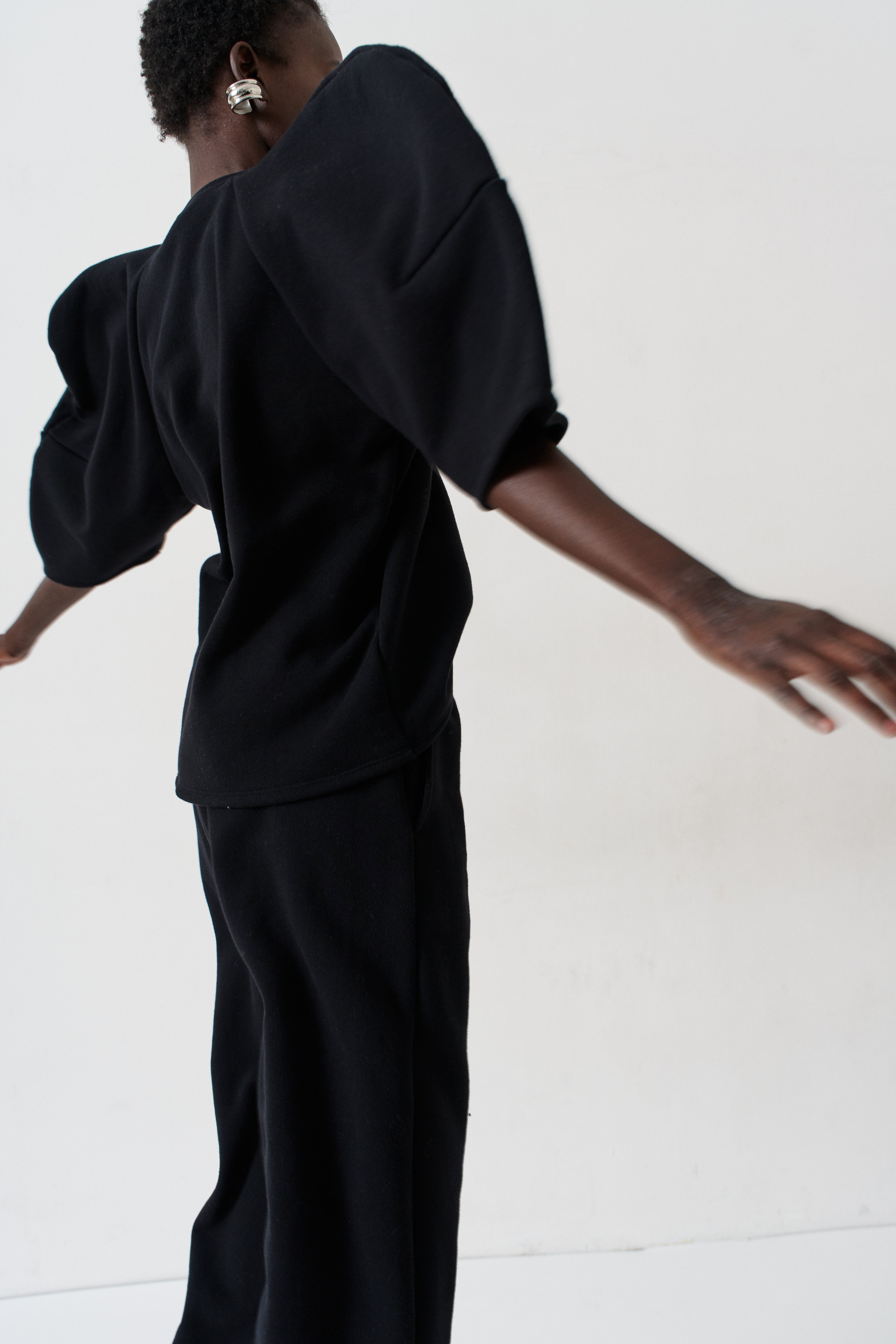 A model from behind wearing a black puff-sleeve top and matching black wide-leg trousers against a plain white studio background.