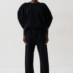 A model stands against a plain white studio background wearing a black two-piece outfit consisting of a voluminous short-sleeve top and matching wide-leg trousers, paired with metallic pointed shoes.