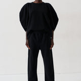 A model stands against a plain white studio background wearing a black two-piece outfit consisting of a voluminous short-sleeve top and matching wide-leg trousers, paired with metallic pointed shoes.