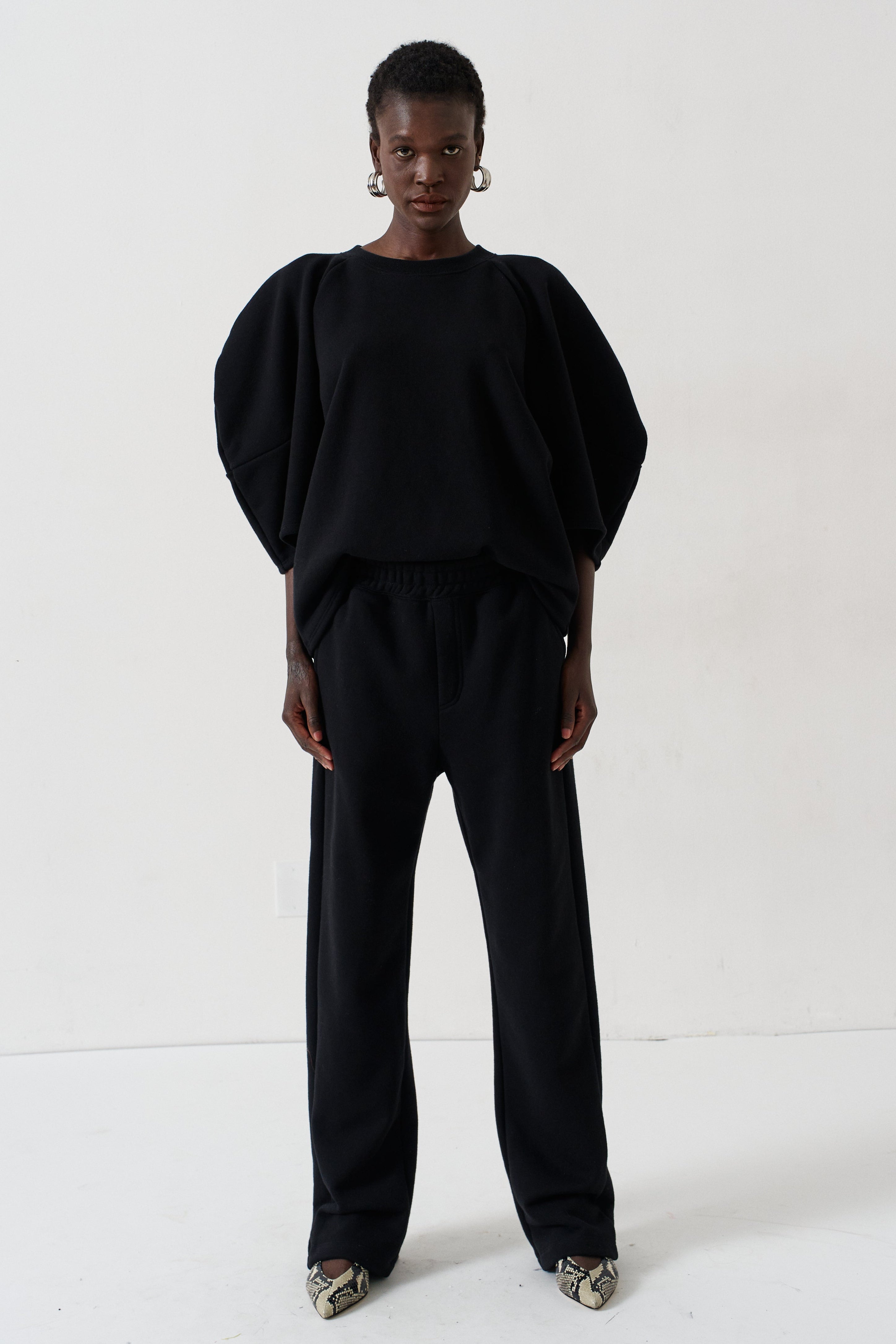 A model stands against a plain white studio background wearing a black two-piece outfit consisting of a voluminous short-sleeve top and matching wide-leg trousers, paired with metallic pointed shoes.