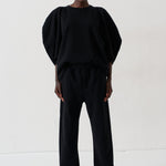 A model stands against a plain white studio background wearing a black two-piece outfit consisting of a voluminous short-sleeve top and matching wide-leg trousers, paired with metallic pointed shoes.