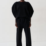 A model stands against a plain white studio background wearing a black two-piece outfit consisting of a voluminous short-sleeve top and matching wide-leg trousers, paired with metallic pointed shoes.