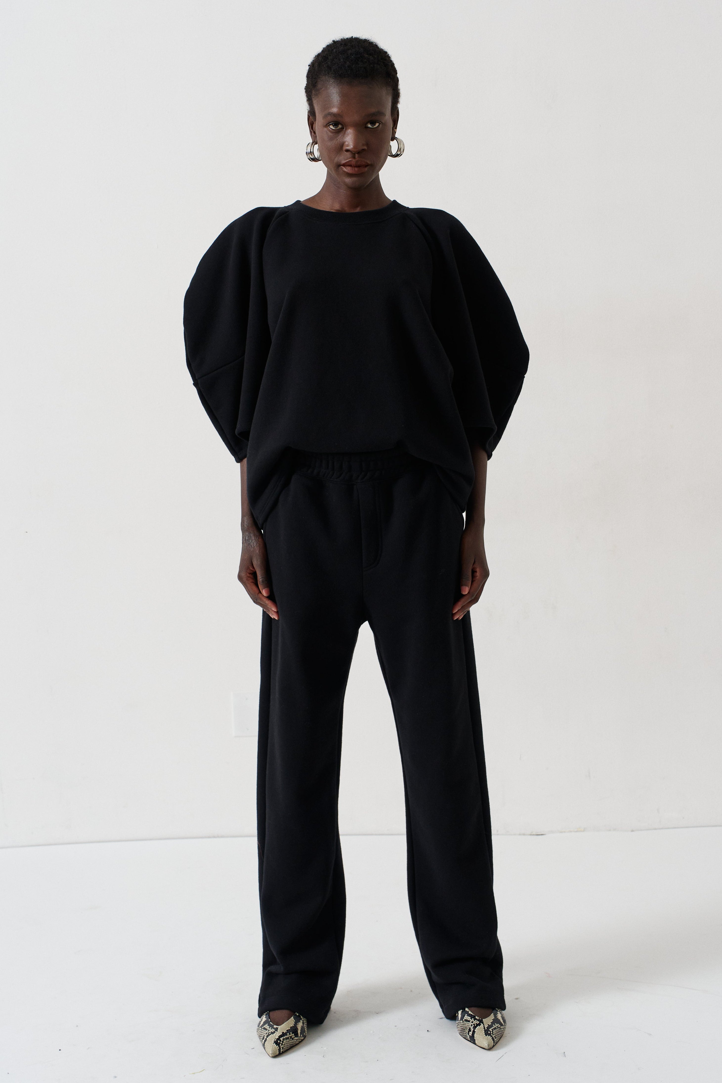 A model stands against a plain white studio background wearing a black two-piece outfit consisting of a voluminous short-sleeve top and matching wide-leg trousers, paired with metallic pointed shoes.