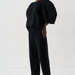 A model wearing a black puff-sleeve top and matching black wide-leg pants, paired with patterned pointed flats, standing against a plain white studio background.