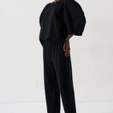 A model wearing a black puff-sleeve top and matching black wide-leg pants, paired with patterned pointed flats, standing against a plain white studio background.
