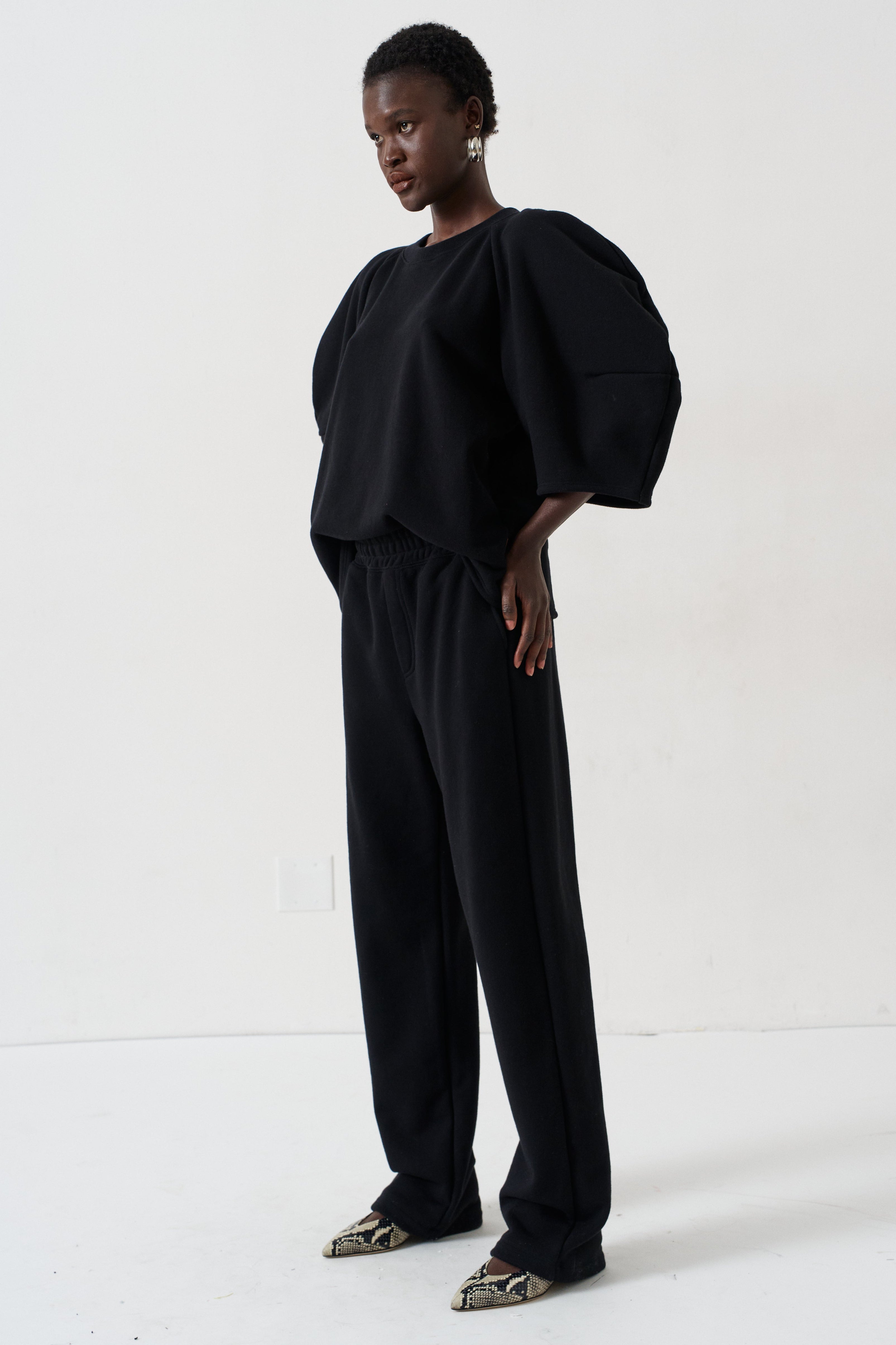 A model wearing a black puff-sleeve top and matching black wide-leg pants, paired with patterned pointed flats, standing against a plain white studio background.