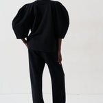 A model viewed from the back wearing a black puff-sleeve top and matching black trousers, standing against a plain white studio background.