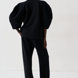 A model viewed from the back wearing a black puff-sleeve top and matching black trousers, standing against a plain white studio background.