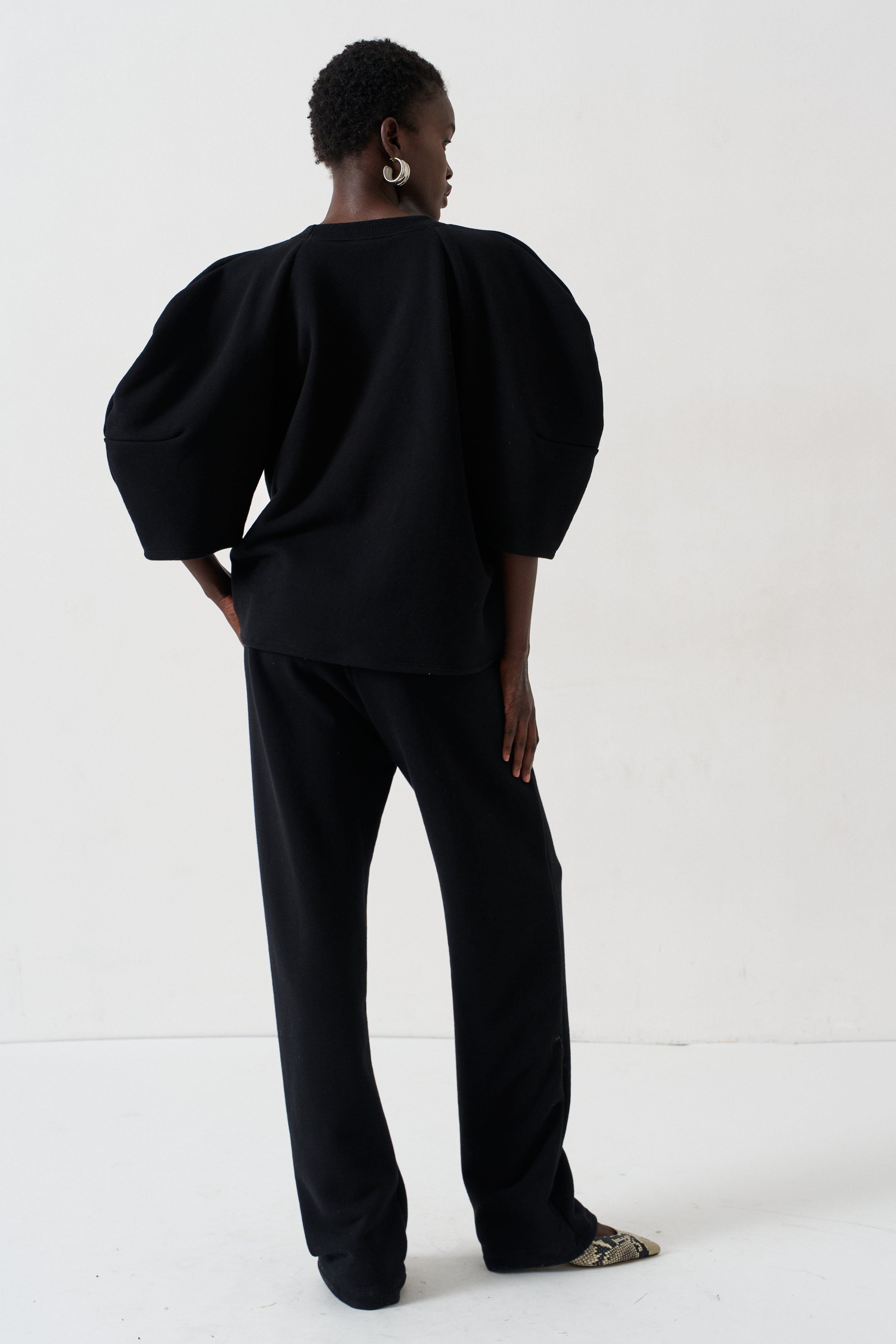 A model viewed from the back wearing a black puff-sleeve top and matching black trousers, standing against a plain white studio background.