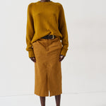 A model stands against a plain white studio background wearing a mustard yellow knit sweater and a matching mustard yellow midi skirt, styled with a black belt and dark pointed-toe heels.