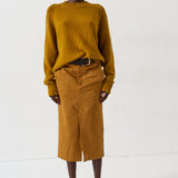 A model stands against a plain white studio background wearing a mustard yellow knit sweater and a matching mustard yellow midi skirt, styled with a black belt and dark pointed-toe heels.