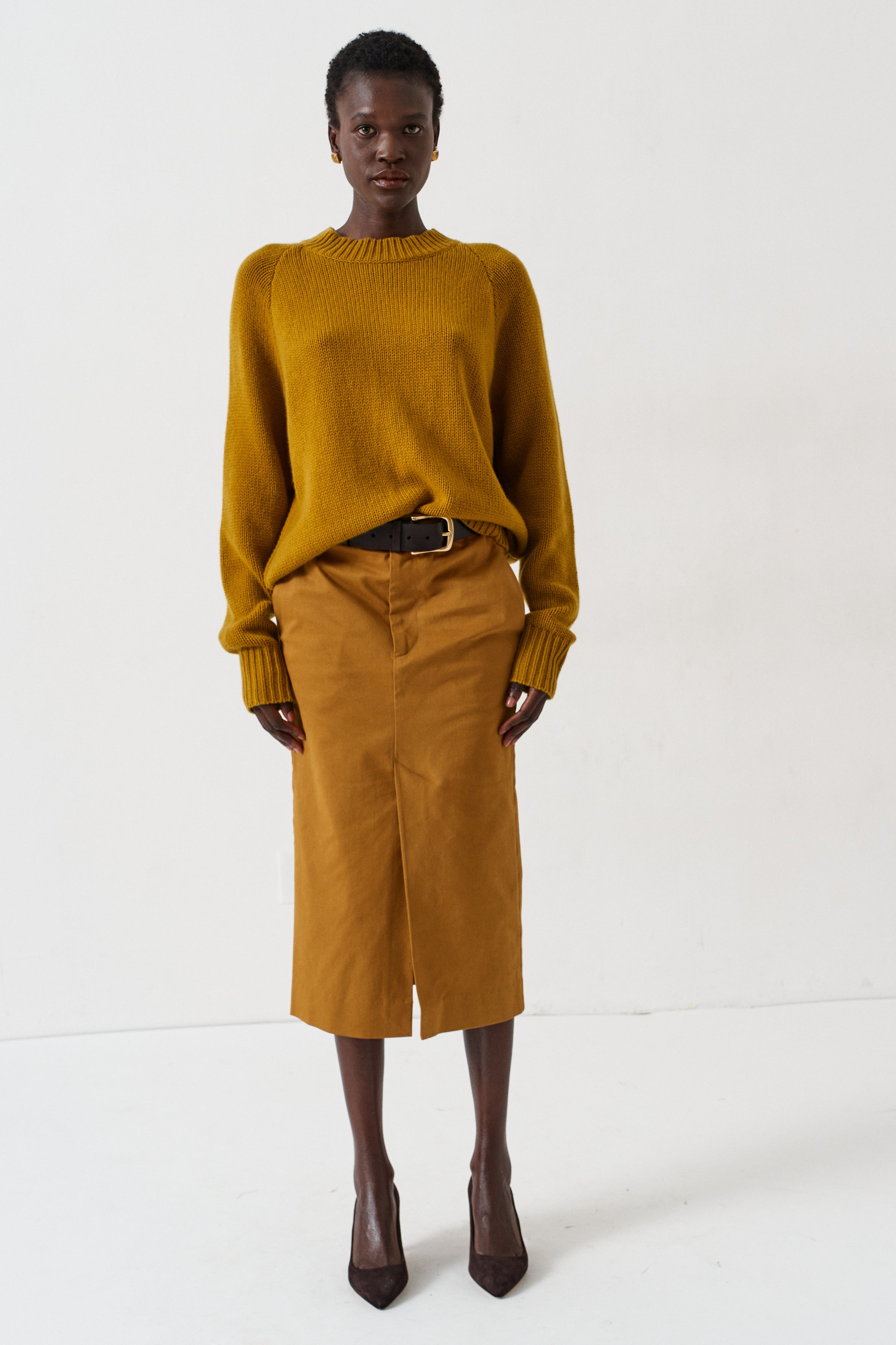 A model stands against a plain white studio background wearing a mustard yellow knit sweater and a matching mustard yellow midi skirt, styled with a black belt and dark pointed-toe heels.