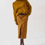 A model wearing a mustard yellow knit mock-neck sweater and a matching mustard yellow midi skirt with a front slit, styled with a dark brown belt and dark brown pointed-toe heels, against a plain white studio background.