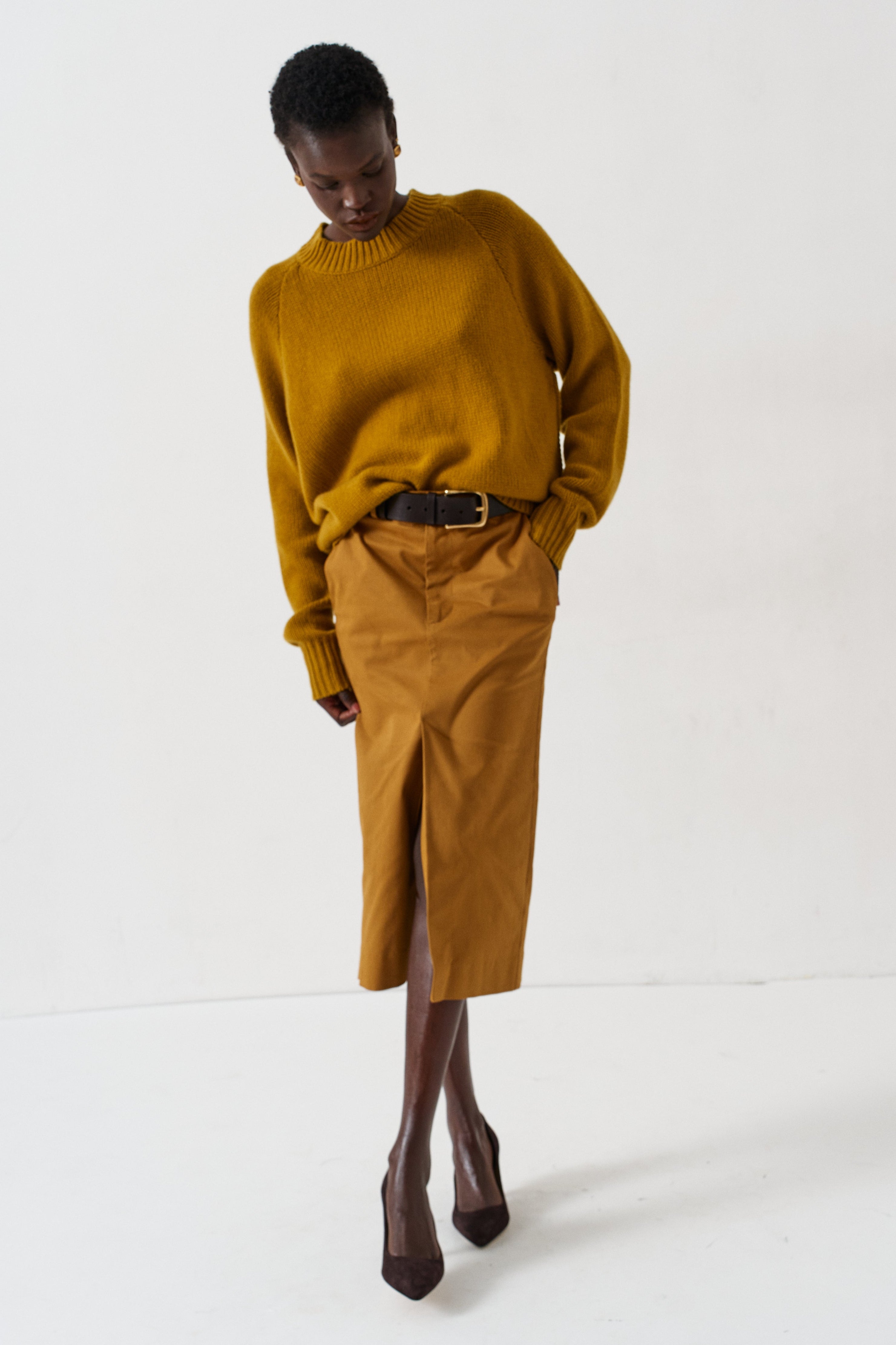 A model wearing a mustard yellow knit mock-neck sweater and a matching mustard yellow midi skirt with a front slit, styled with a dark brown belt and dark brown pointed-toe heels, against a plain white studio background.