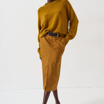 A model stands against a plain white studio background wearing a mustard yellow ribbed knit sweater and matching mustard yellow midi skirt with a black belt, paired with dark brown pointed-toe heels.