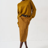 A model stands against a plain white studio background wearing a mustard yellow ribbed knit sweater and matching mustard yellow midi skirt with a black belt, paired with dark brown pointed-toe heels.