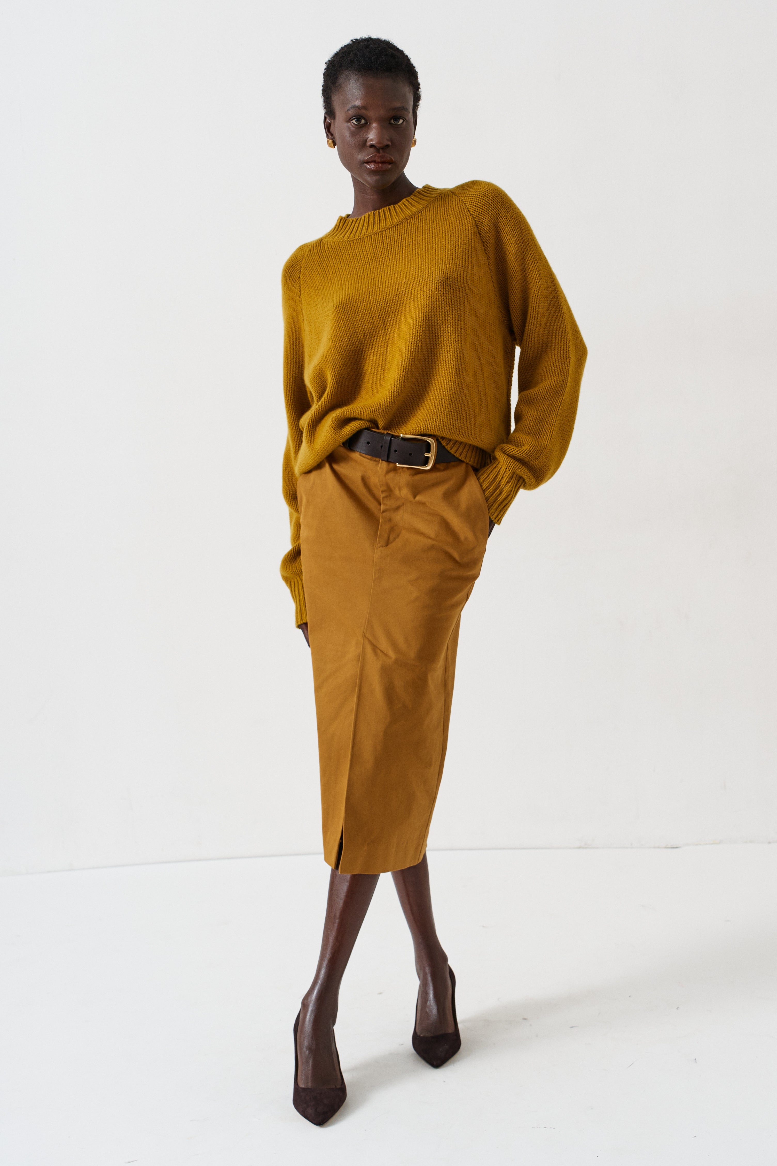 A model stands against a plain white studio background wearing a mustard yellow ribbed knit sweater and matching mustard yellow midi skirt with a black belt, paired with dark brown pointed-toe heels.